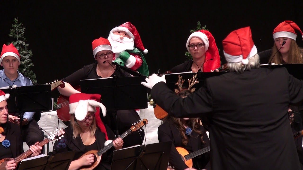 Mladi člani orkestra v božičnih kapah igrajo tamburice, dirigent jih vodi.